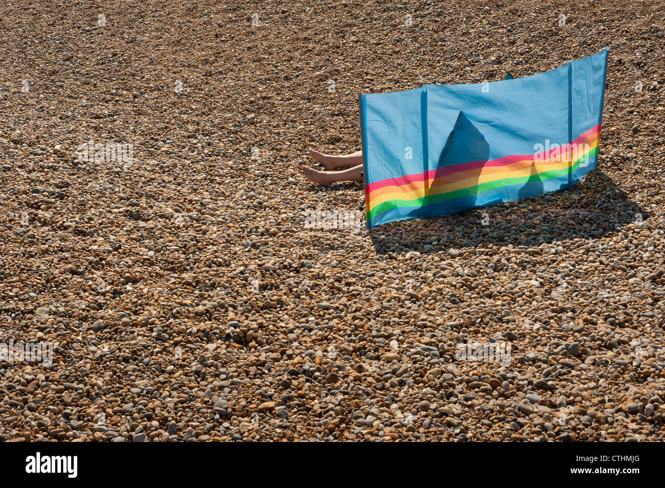Obscured sun bather lying on a pebble beach and sheltering behind a colourful windbreak Stock Photo