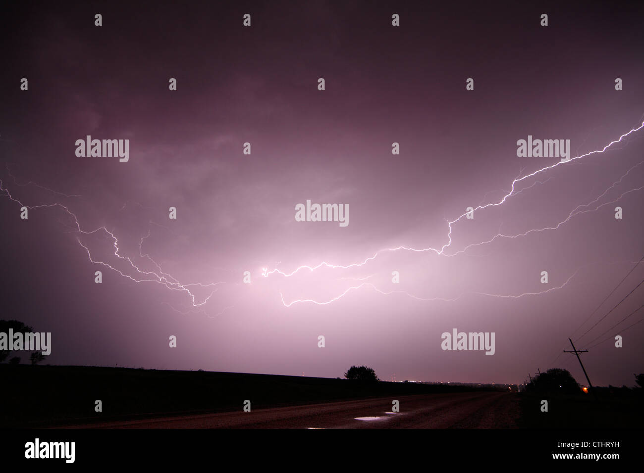 Multiple lightning bolts crawl along the underside of a severe thunderstorm over a wet gravel road at night. Stock Photo