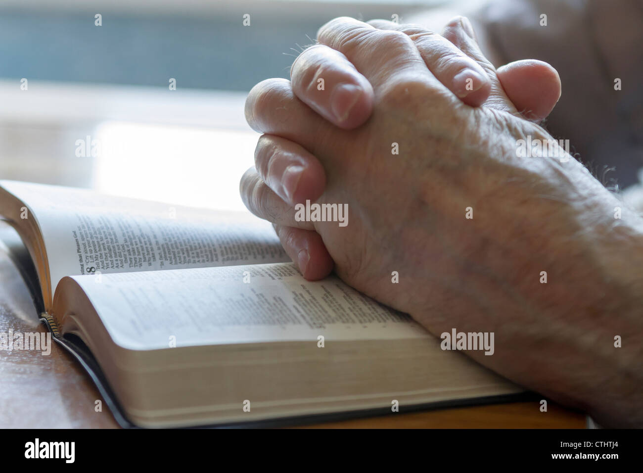 weathered old man's hands clasped in prayer over open Bible Stock Photo