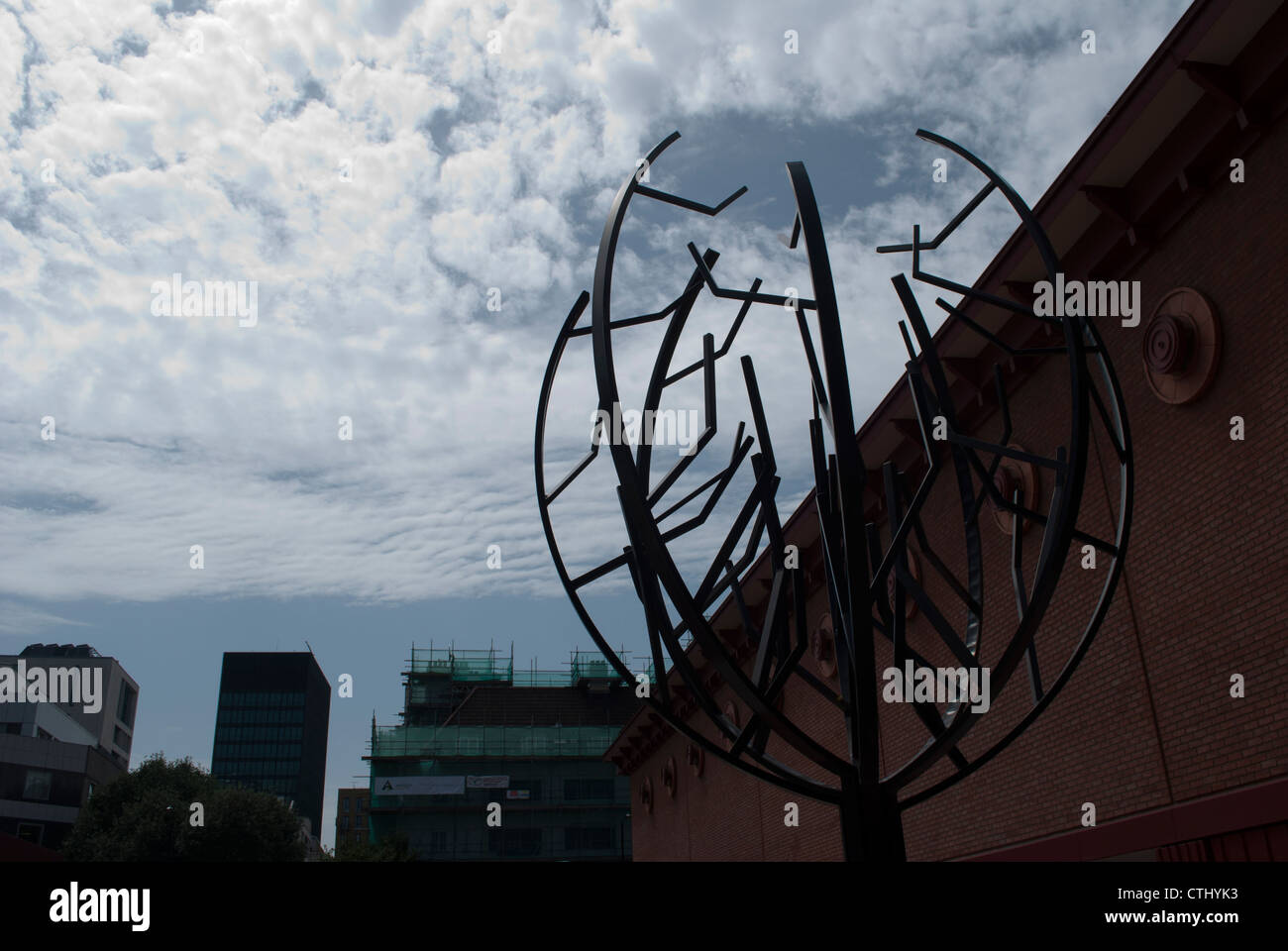 Metal tree sculpture silhouetted against cloudy sky and red brick wall of British Library Stock Photo