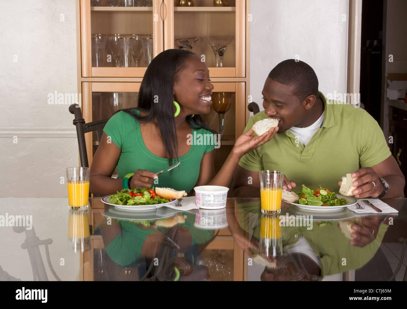 Young black African American couple sitting by glass table and eating meal of salad, bagels with cream cheese and orange juice Stock Photo