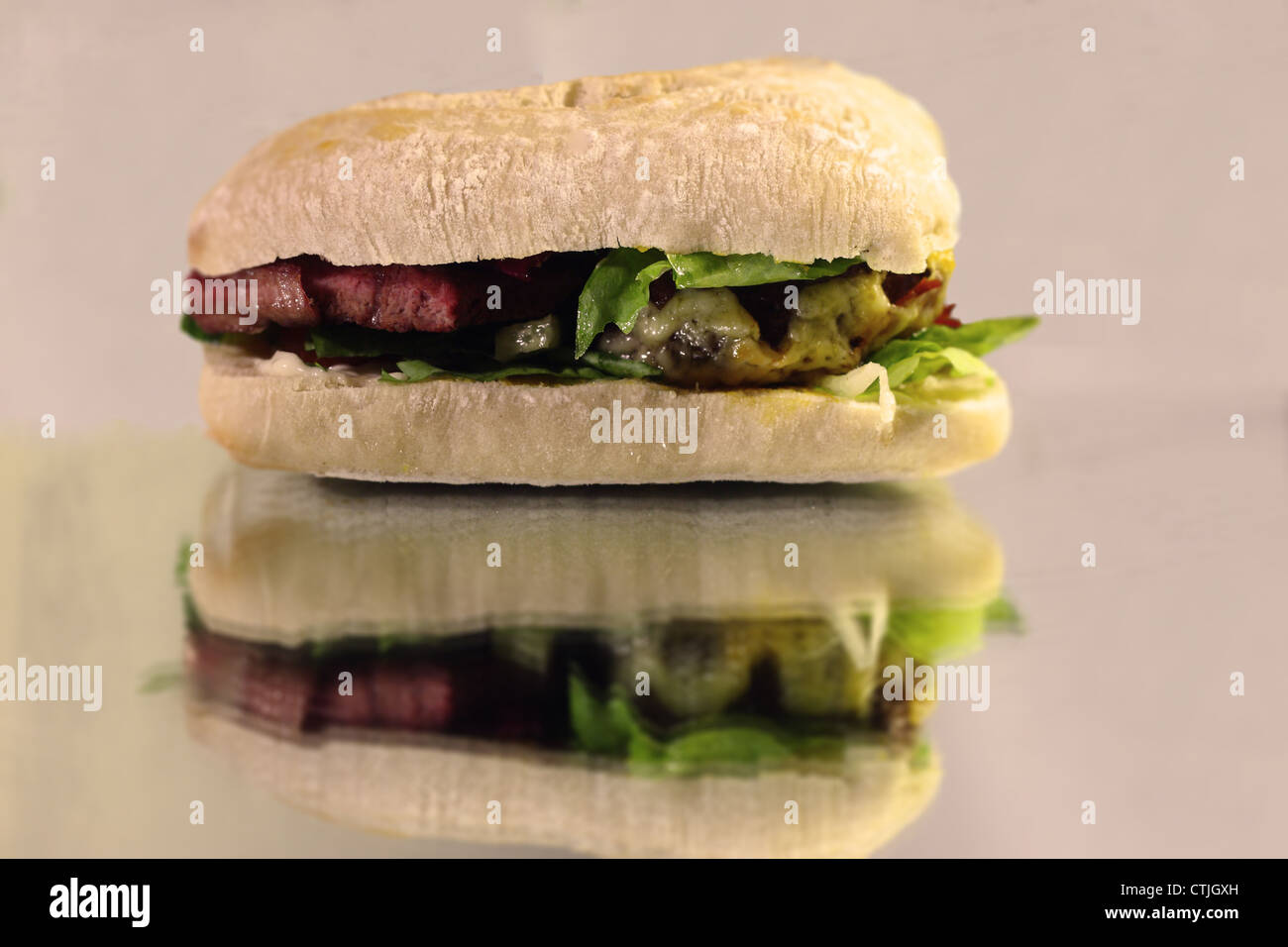 A floury burger bap containing a beef patty and a slice of steak covered with garnishes sitting on top of a reflective surface. Stock Photo