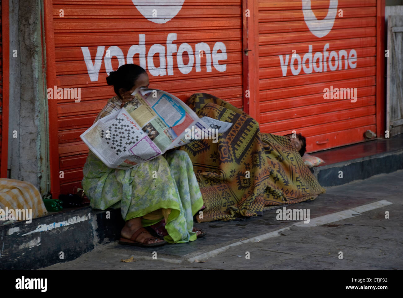 Homeless woman reading newspaper with her partner sleeping aside. Street scene - Mumbai, India Stock Photo