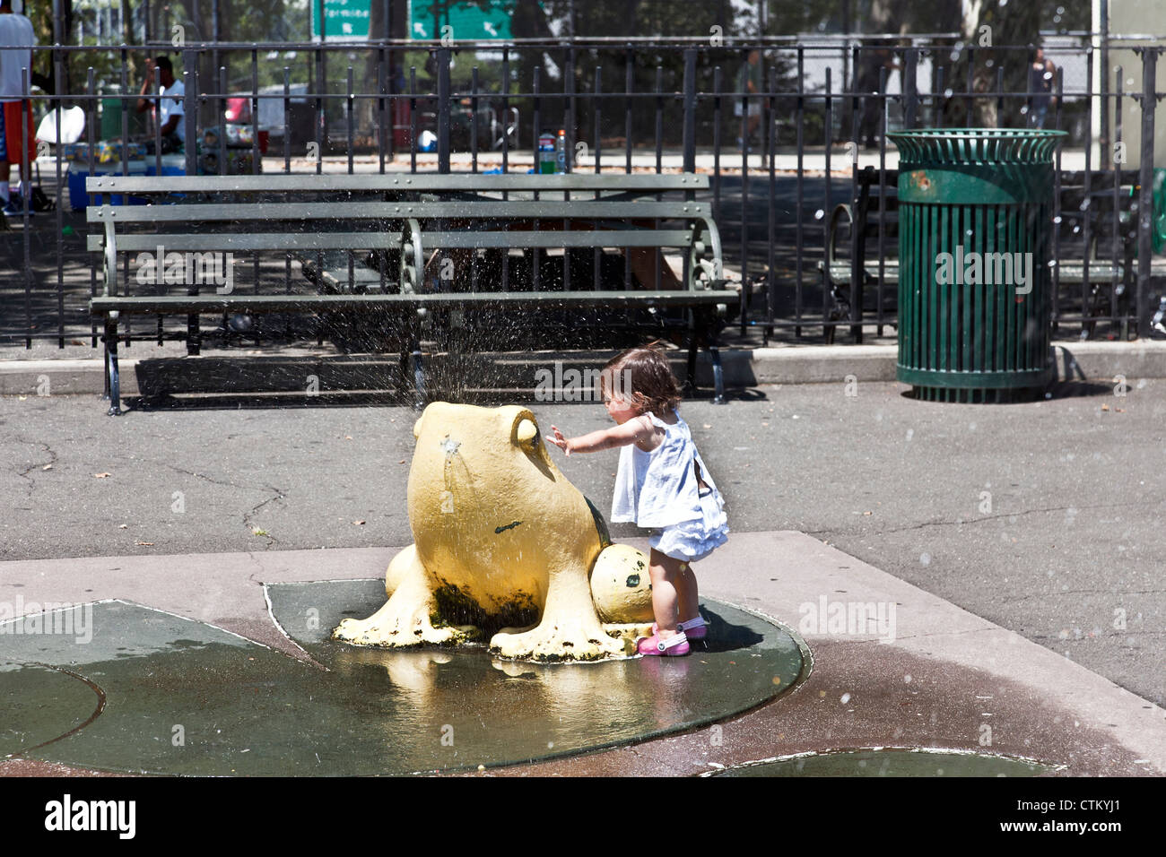 tiny girl toddler in pink crocs & ruffles explores yellow frog fountain on sunny summer day in DeWitt Clinton park Manhattan Stock Photo
