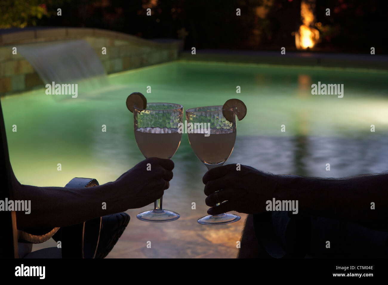 Couple Sitting At Pool Side At Night With Two Glasses Together With Lime Wedges And Pool Lights; Palm Springs, California, USA Stock Photo