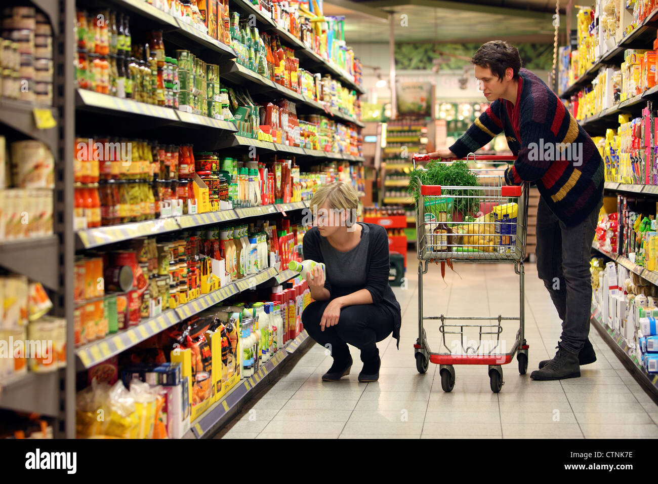 A young couple in a large supermarket, shopping with a trolley, walking through the aisles with shelfs full of food products. Stock Photo