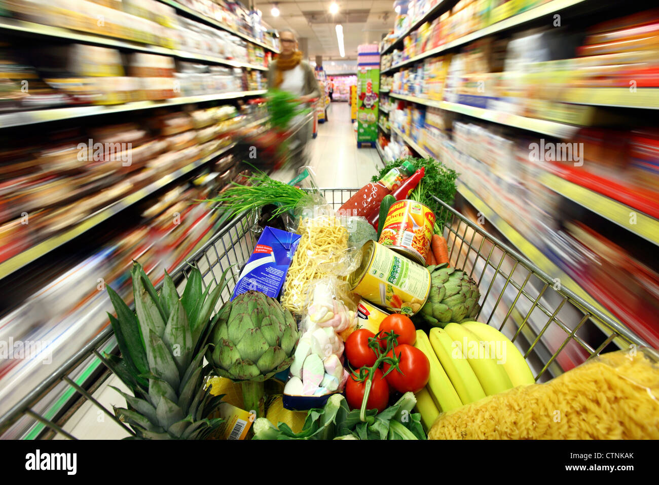 Food hall, full shopping trolley is pushed through a hallway with several food products. Large supermarket, self service. Stock Photo