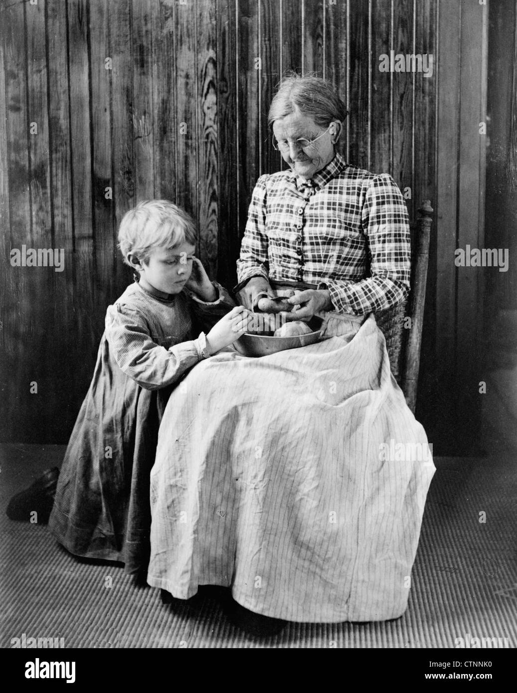 Anticipation - an elderly woman seated in a chair peeling potatoes with a young girl, possibly a granddaughter, kneeling on the floor next to her, watching, circa 1897 Stock Photo