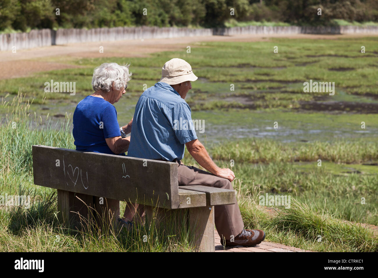 elderly man and woman sat on bench in the sunshine at Langston harbour at low tide with I Love you grafitti Stock Photo