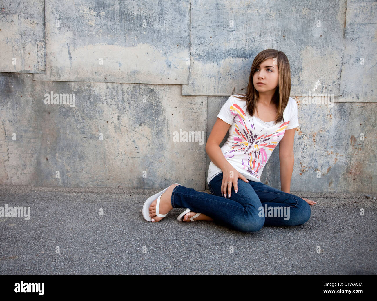 Outdoor photo of pretty teenage girl seated on pavement against grunge wall. Stock Photo