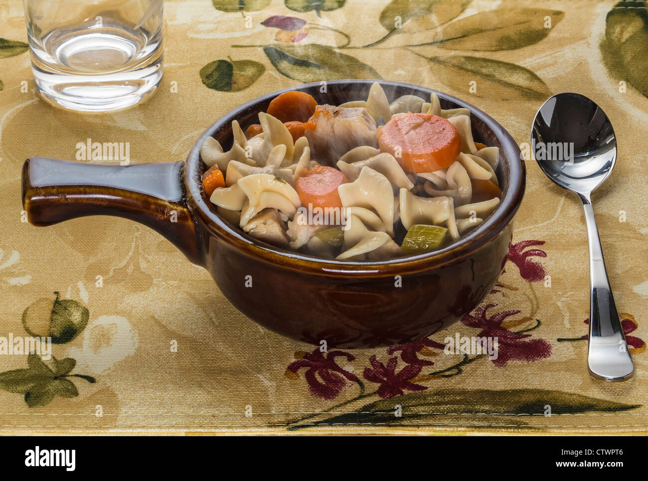 stoneware bowl of chicken noodle soup on woven placemat with spoon and water glass Stock Photo
