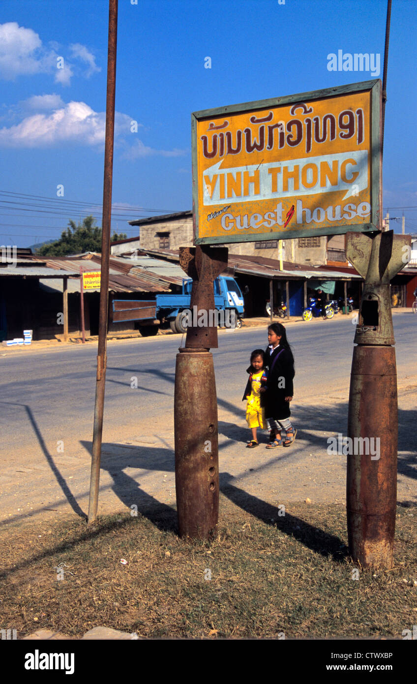 Two Laotian Girls or Sisters Walk Past Guest House or Hotel Sign Supported on Bomb Casings of Recycled US Cluster Bombs Used in Vietnam War Phonsavan Laos Stock Photo