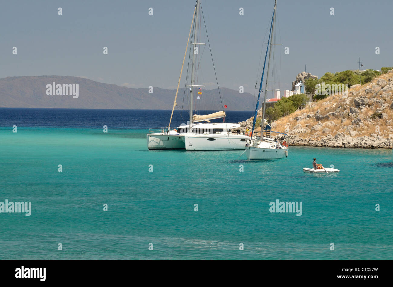 A catamaran, a sailboat and a raft crowd the amazing bay at Aghia Marina with the turkish peninsula of Datca in the background. Stock Photo