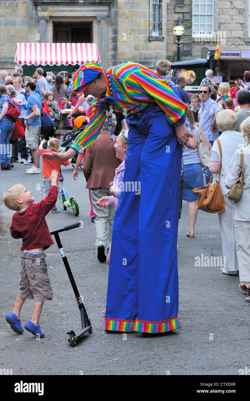 A man dressed as a clown walks on stilts and gives high five to young boy with scooter. Pollok Park family day. Stock Photo