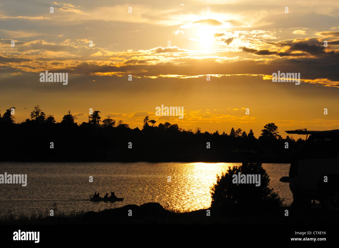 Silhouette of a boat of trout fishermen going down a lake as the sun sets with people camping nearby Stock Photo