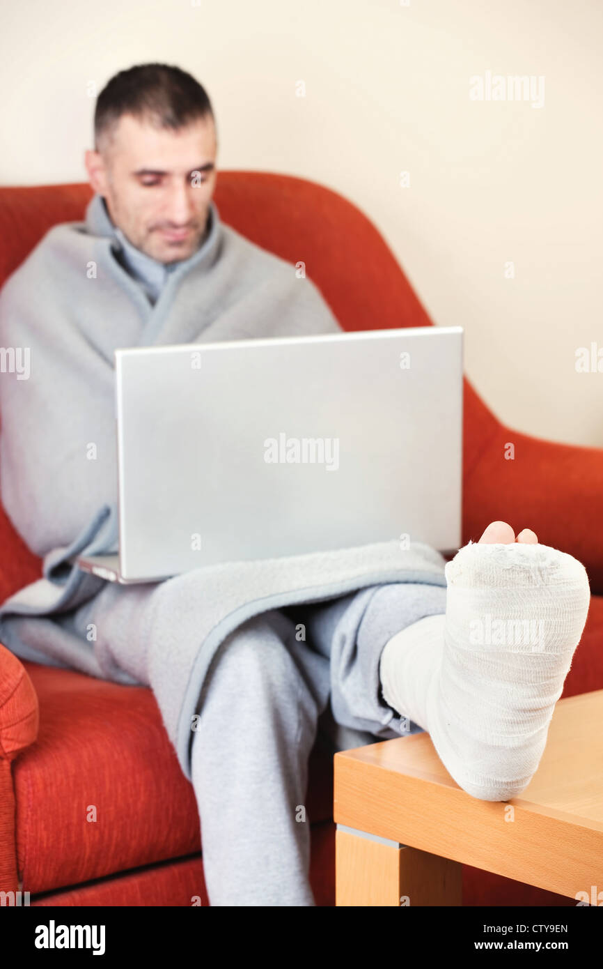 man with a broken leg on a sofa at home working on laptop Stock Photo