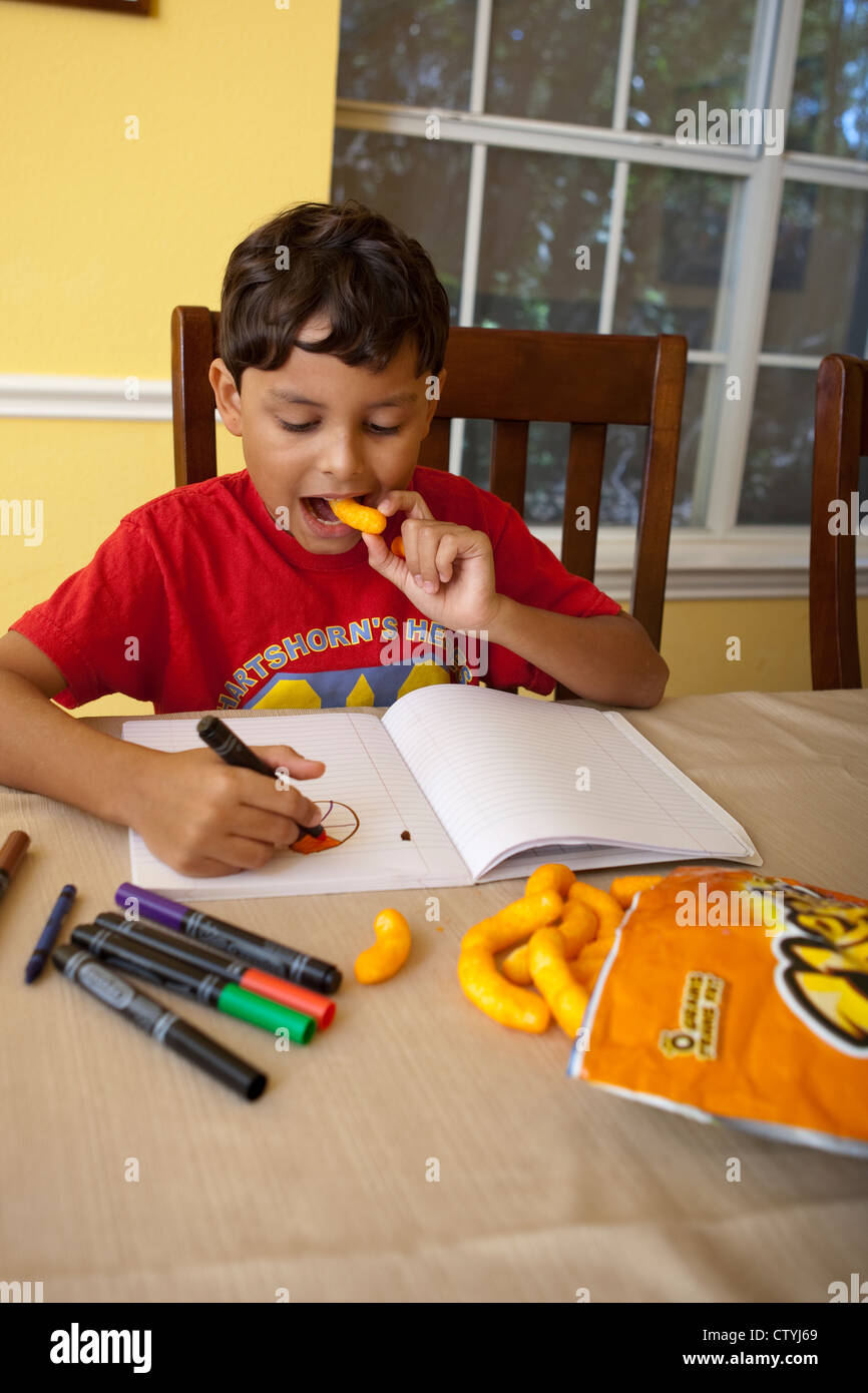 7 and 8 year old Mexican-American boy snacks on junk food, Cheetos, while drawing at home with markers. Stock Photo