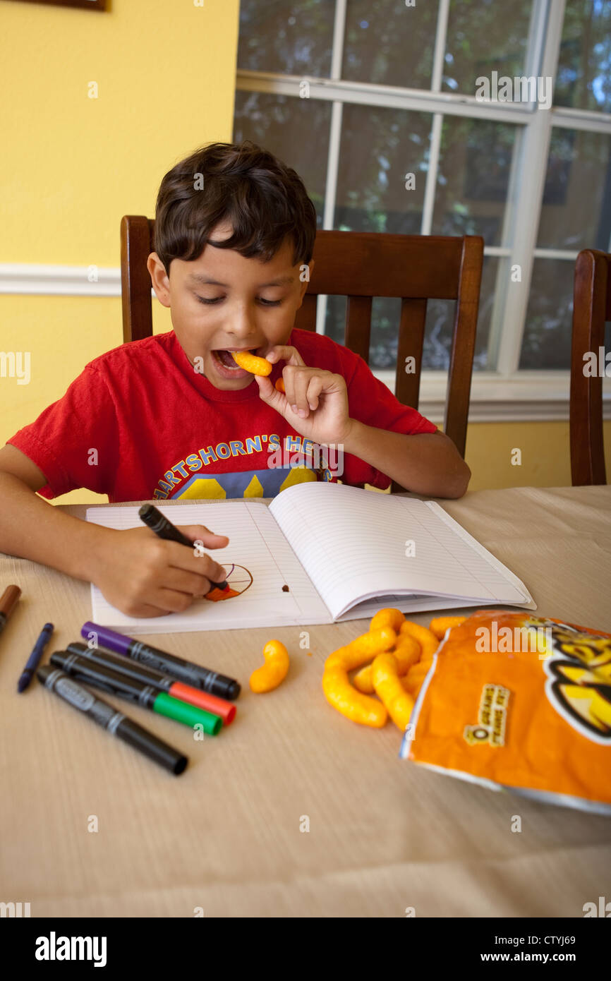 Eight-year-old Mexican-American boy snacks on junk food, Cheetos, while drawing at home with markers. Stock Photo