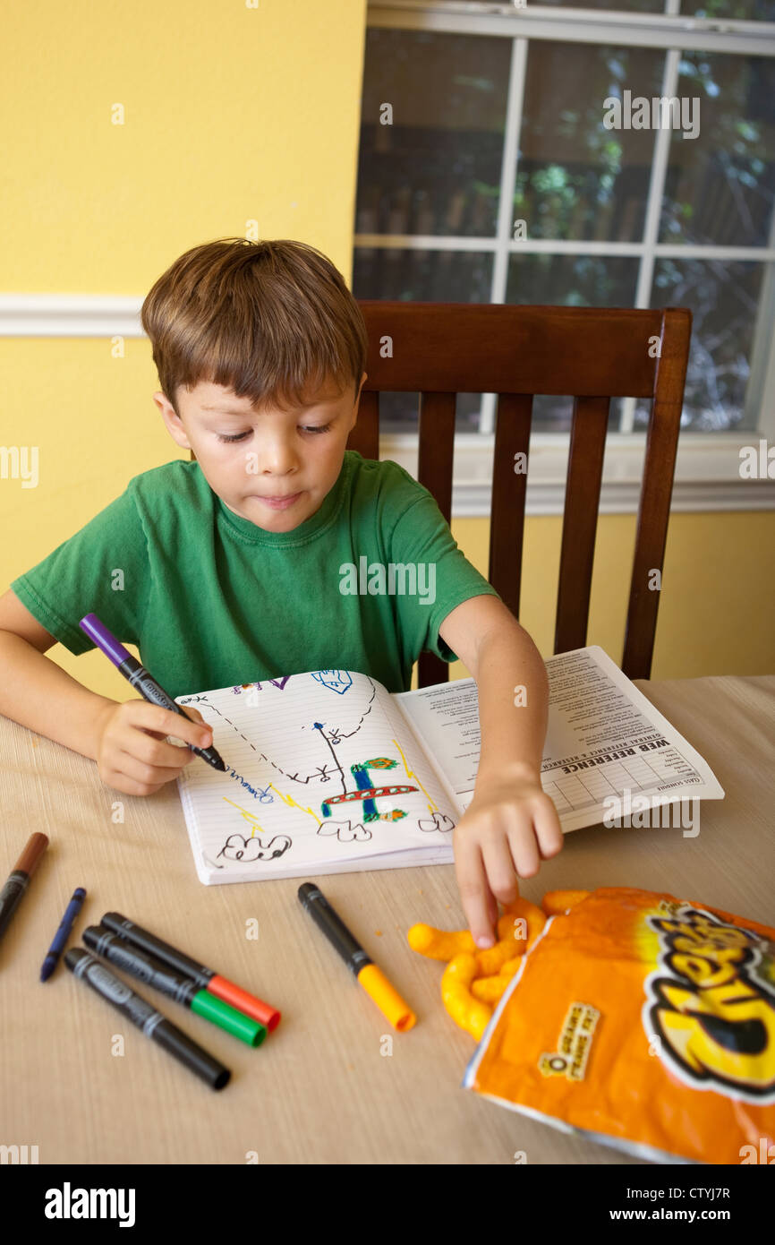 Seven-year-old Mexican-American boy snacks on junk food, Cheetos, while drawing at home with markers. Stock Photo