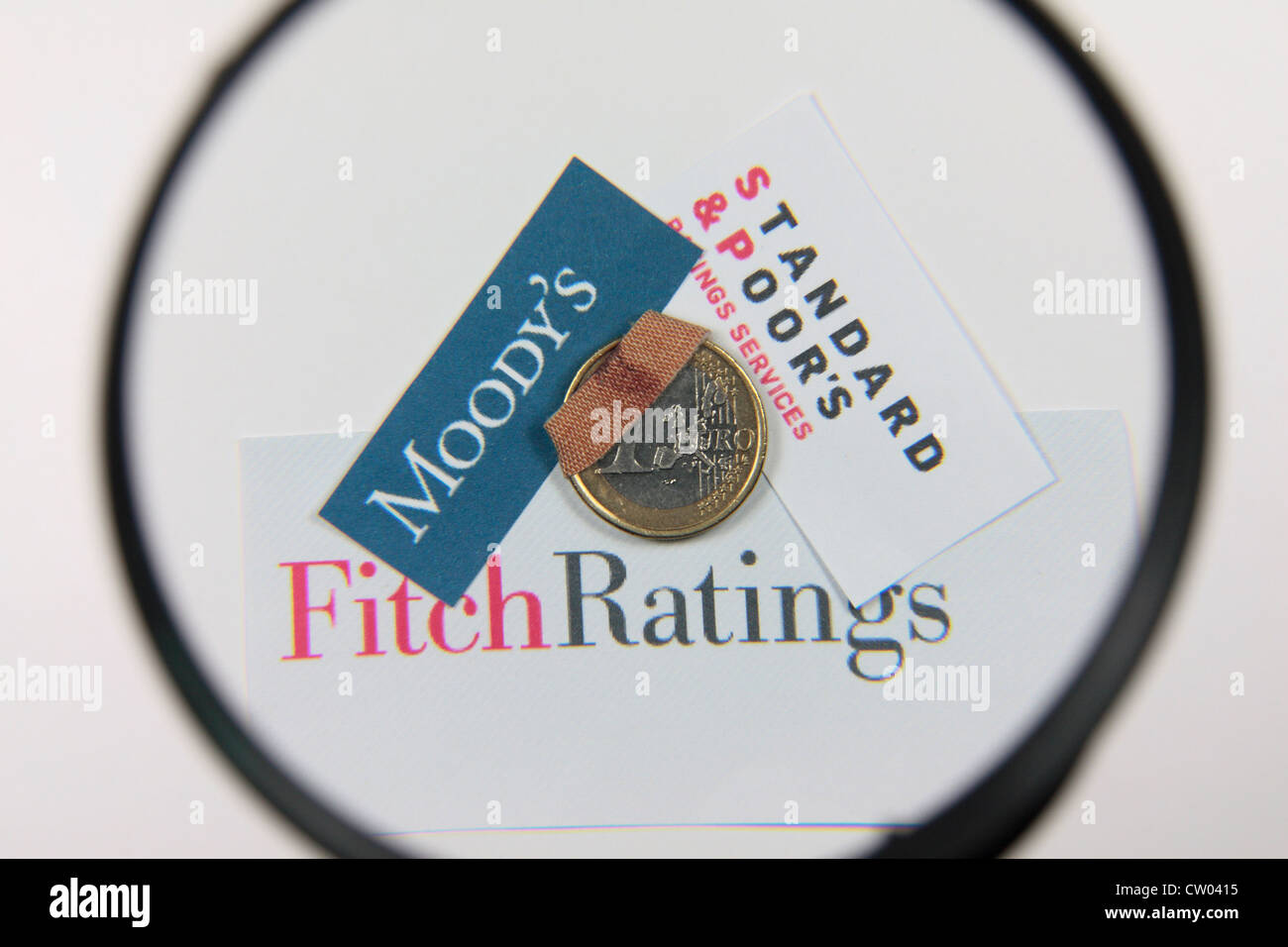An 'injured', 'damaged' or 'wounded' one Euro coin under a magnifying glass with the logos for the three credit rating agencies. Stock Photo