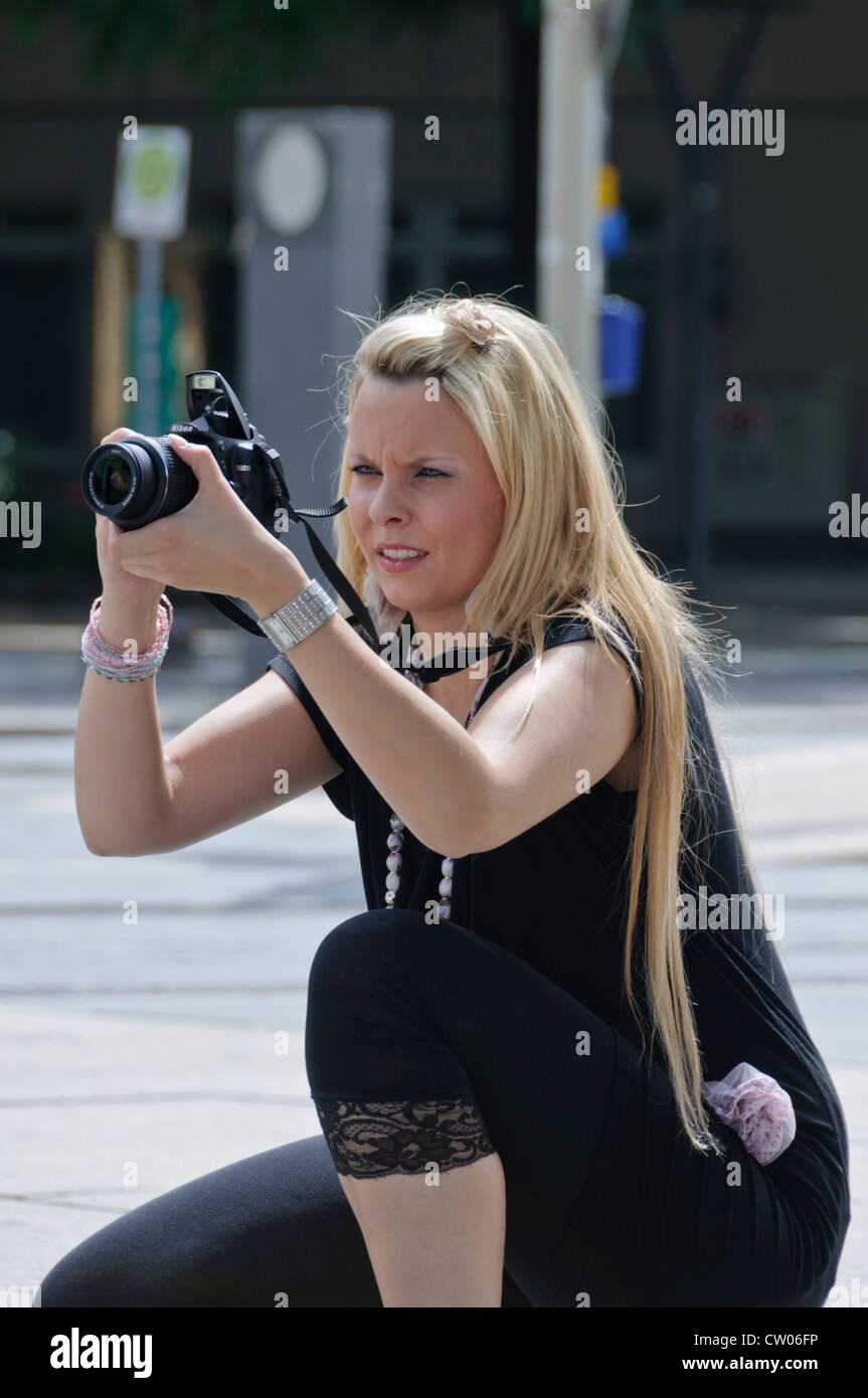 Beautiful young female photographer with long blond hair and black dress taking pictures with a NIKON D5000 DSLR 18-55 mm lense Stock Photo