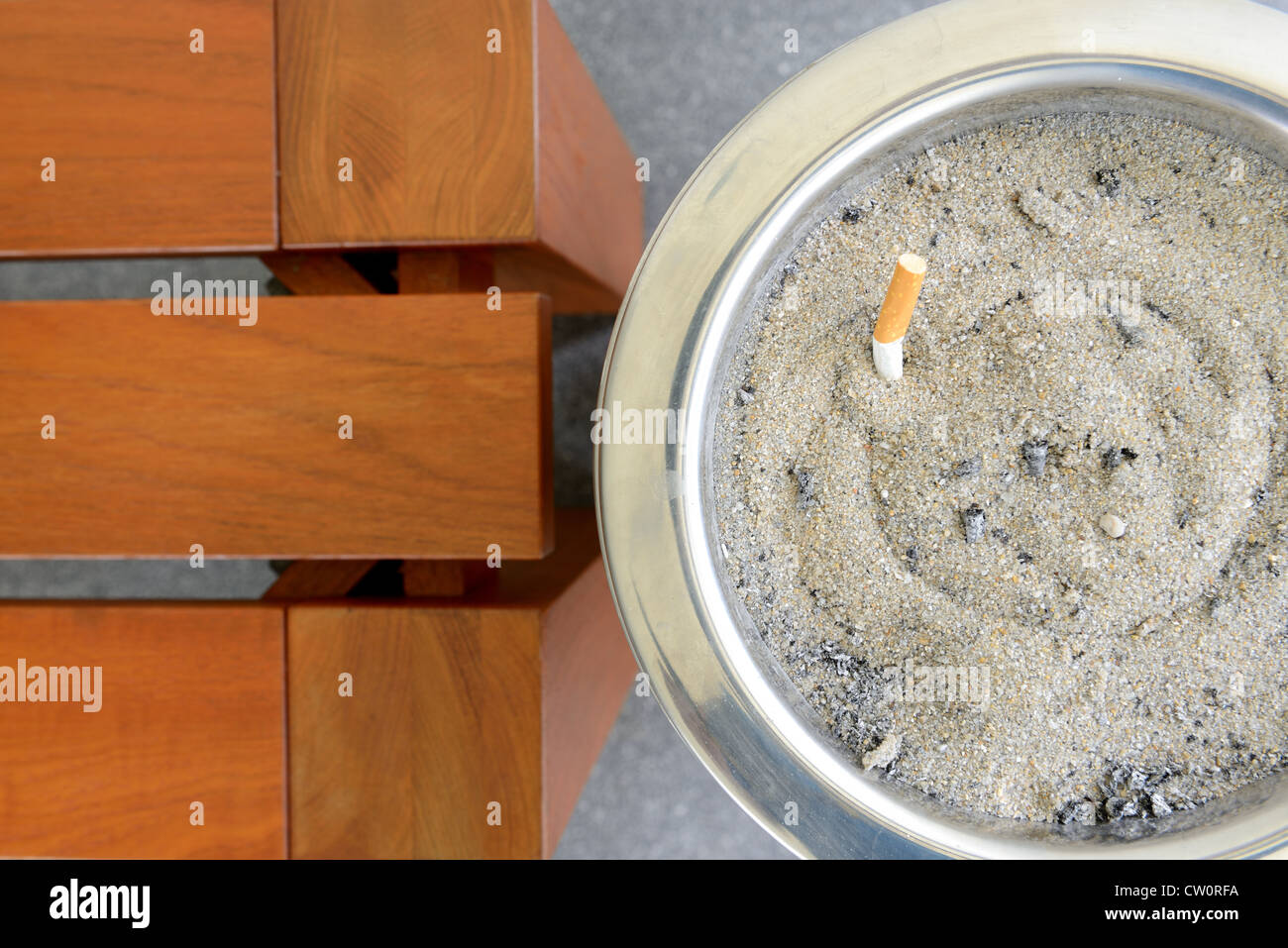 Cigarette in sand ashtray bin is next to wooden bench. Stock Photo