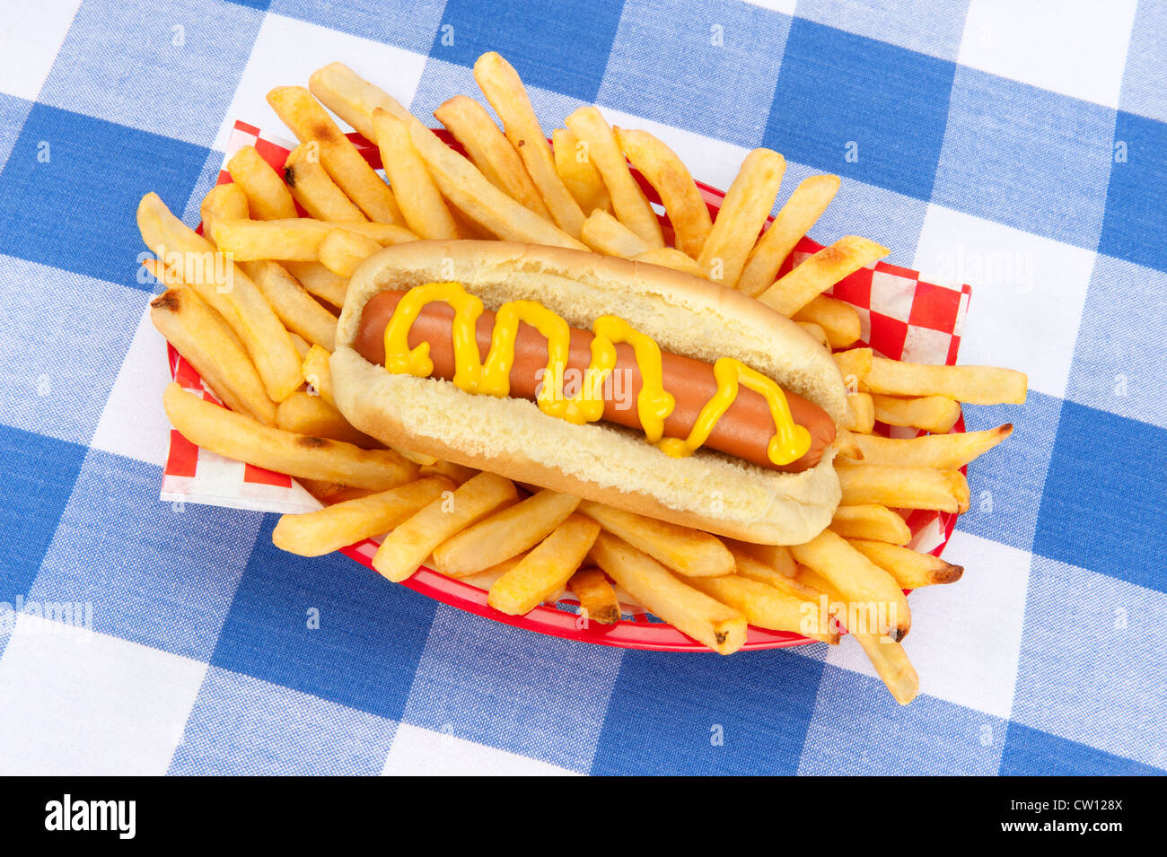A basket of french fries and a hotdog with mustard on a tabletop covered with a checkered tablecloth. Stock Photo