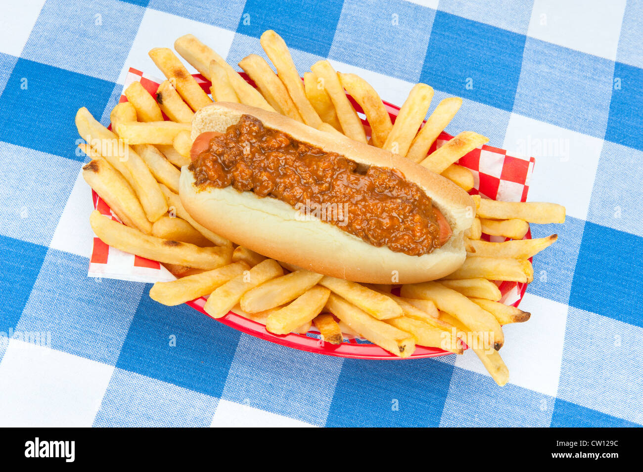 A fresh chilidog with a basket of French fried potatoes on a blue, checkered tablecloth. Stock Photo
