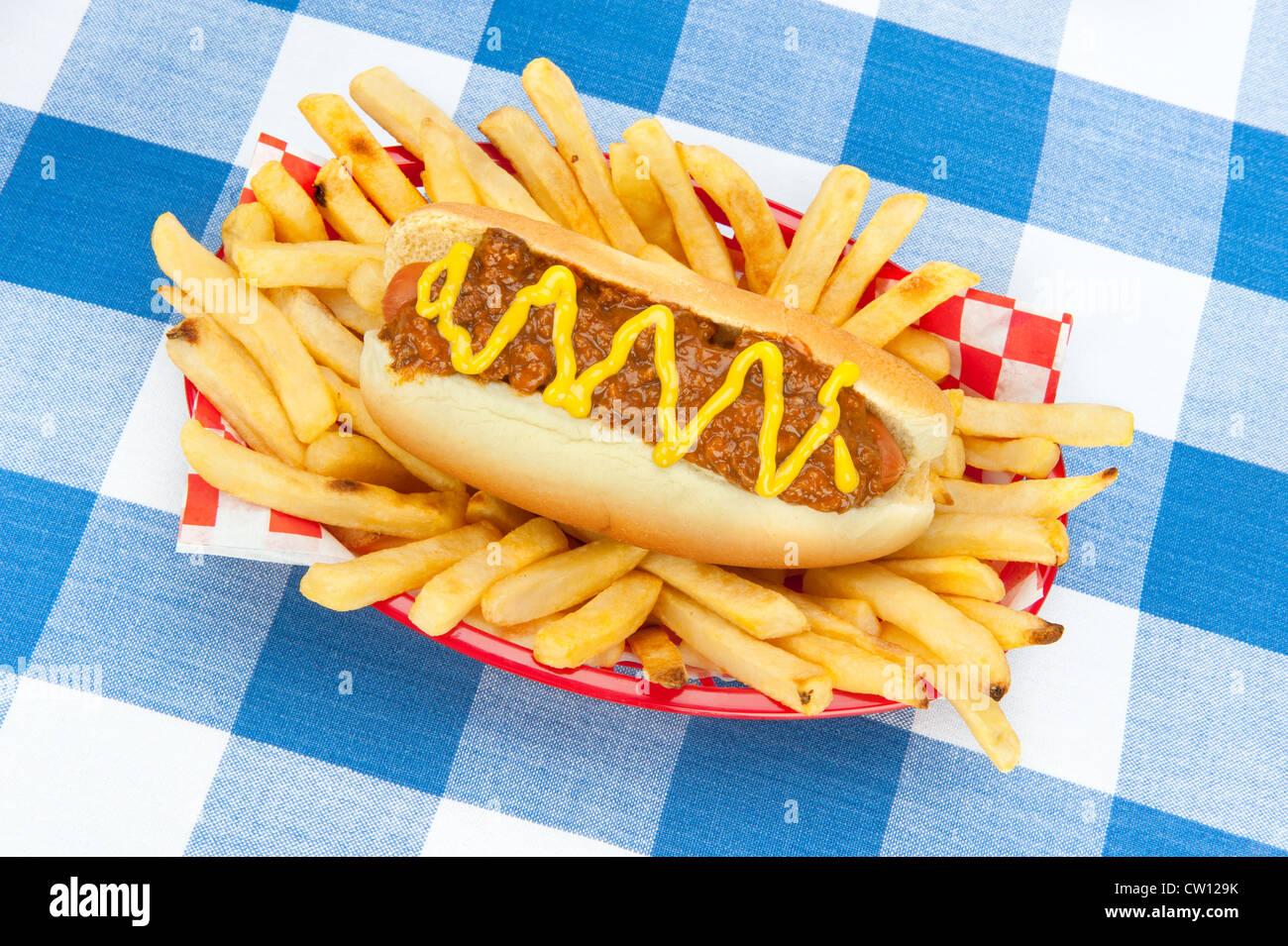 A chilidog with mustard in a basket of fresh French fries on a blue and white checkered tablecloth. Stock Photo