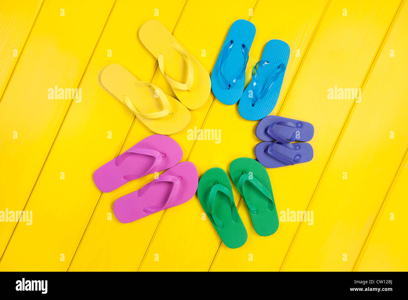 An assortment of colorful rubber flip flops in a circular pattern on a wooden, yellow pool deck. Stock Photo