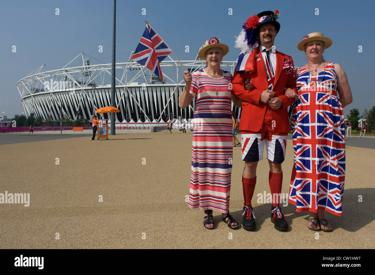 A tall son in the middle accompanies his mother (l) and sister (r) in the Olympic Park during the London 2012 Olympics. Wearing Union Jack designs dresses and shorts plus a tie and flag to wave, the trio stand in front of the main Olympic stadium before the large crowds arrive later in the day. They have tickets to watch the Hockey and stand smiling as eccentric Brits wearing their national colours. Stock Photo