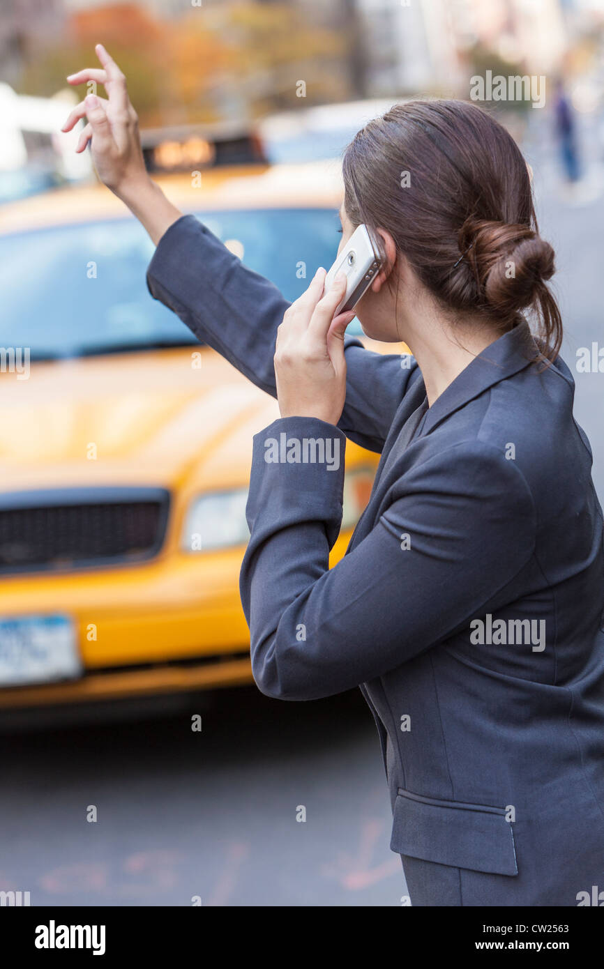 A young woman or businesswoman hailing a yellow Taxi cab while talking on her cell phone in a modern city Stock Photo