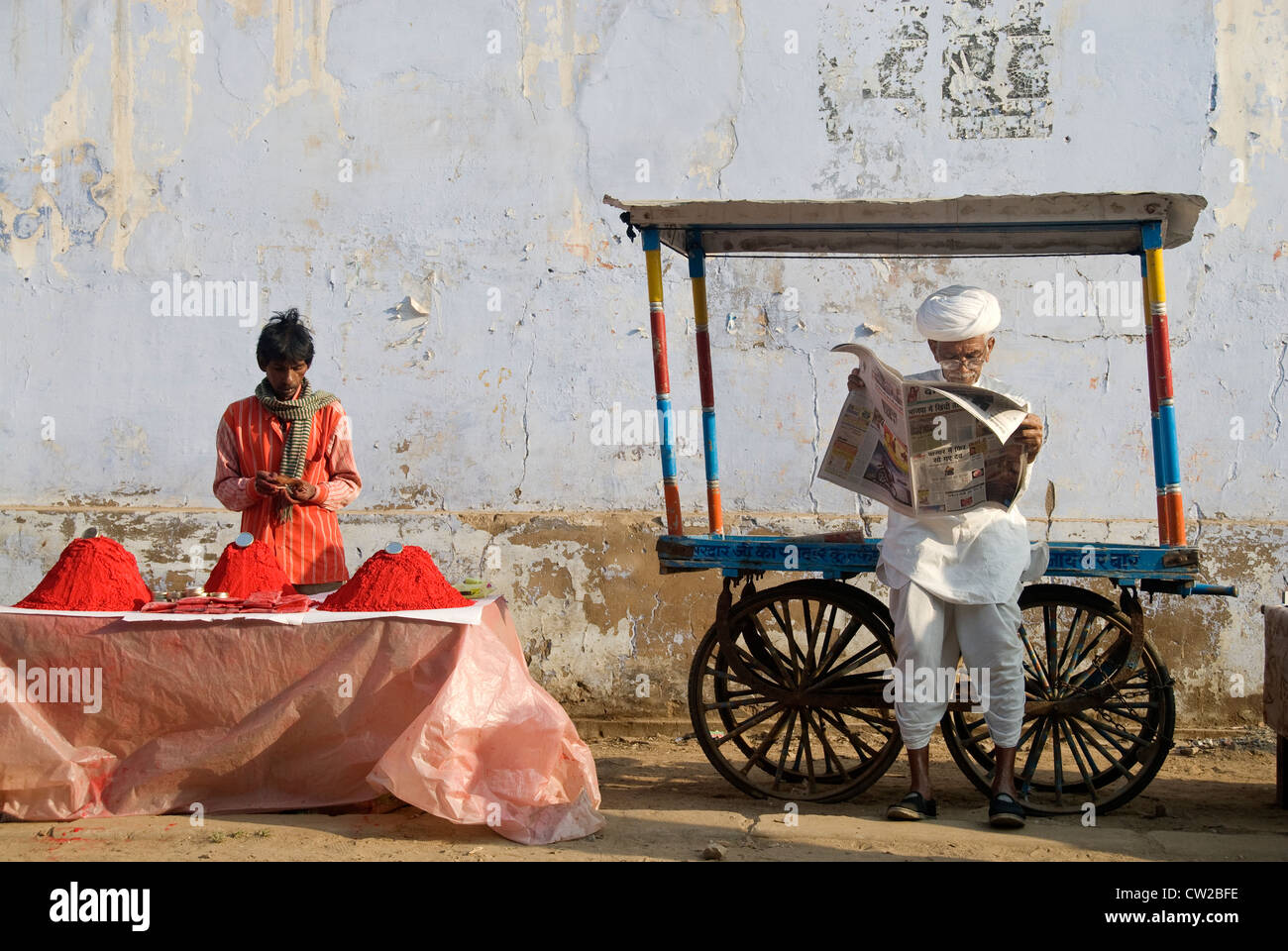 Elderly man reading newspaper. Wayside vendor selling color dyes beside him - Pushkar, Rajasthan Stock Photo