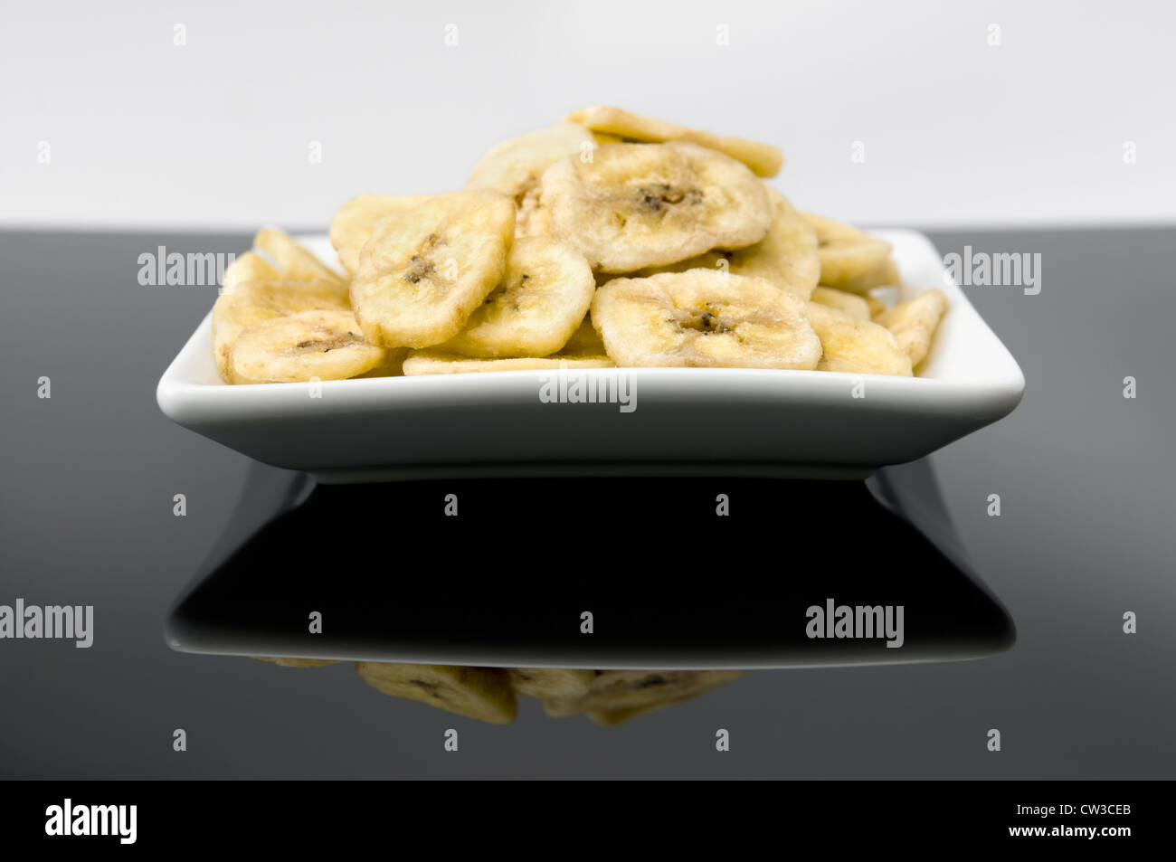 Dried banana chips in white bowl in black reflective surface against a white background Stock Photo