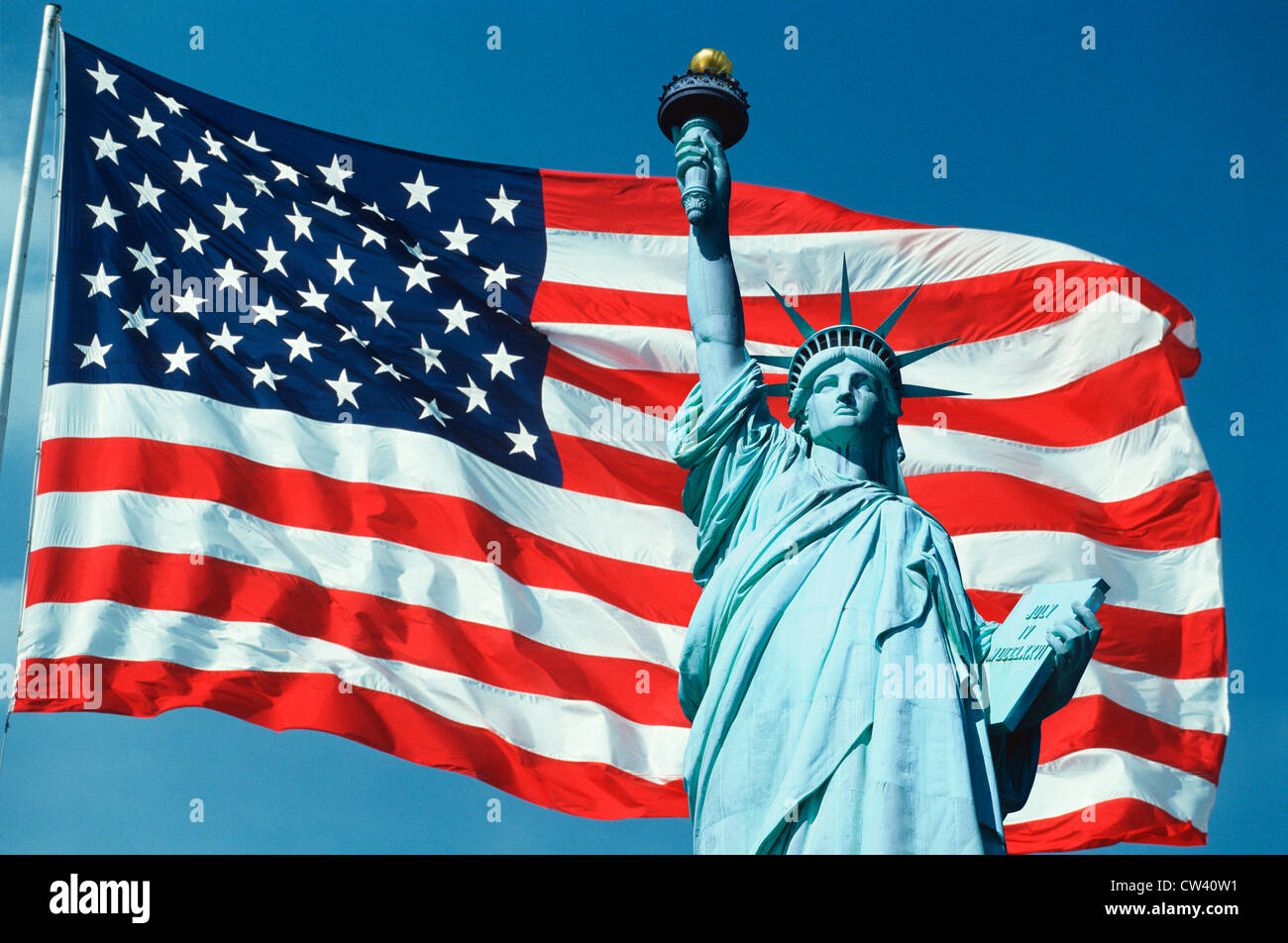 This an American flag on flagpole waving in wind set against blue sky. top half Statue Liberty digitally composited onto right Stock Photo