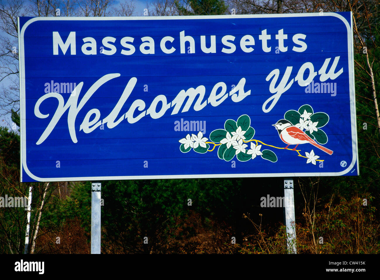 This is a road sign that says Massachusetts welcomes you. It is against a blue sky. On the sign are the state bird and flowers. Stock Photo