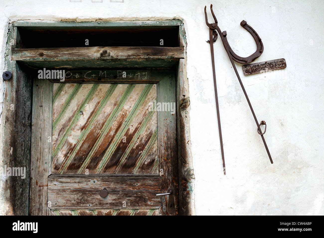 Horse shoe and Blacksmiths metal tongues mounted onto a white washed farm wall next to a paneled wooden door. Stock Photo