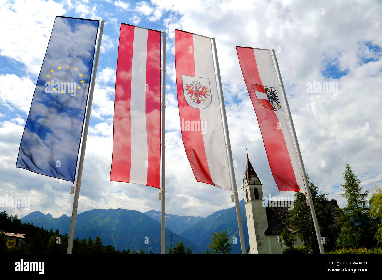 European, Austrian, regional and local flags of Austria in full display and traditional Austrian spired church in the background Stock Photo