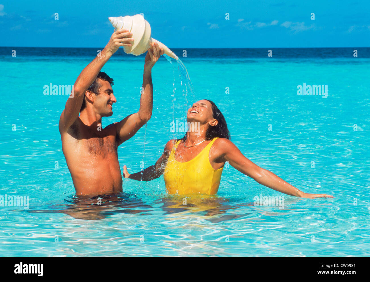 Couple chilling out pouring water from conch shell onto each other in aqua marine waters during island holiday Stock Photo