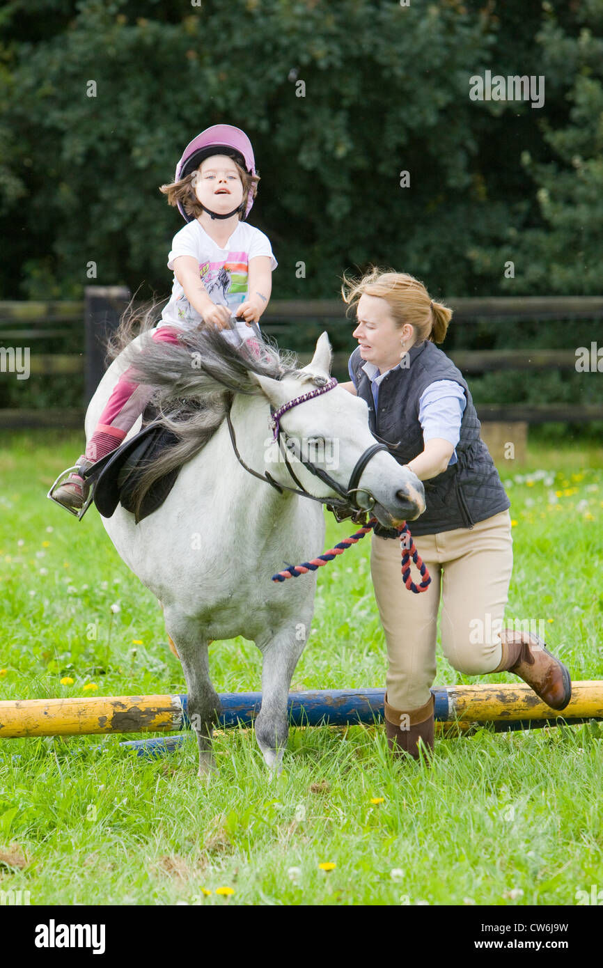A young girl on a white pony being lead over a small wooden jump in a grass paddock in the English countryside in summer Stock Photo