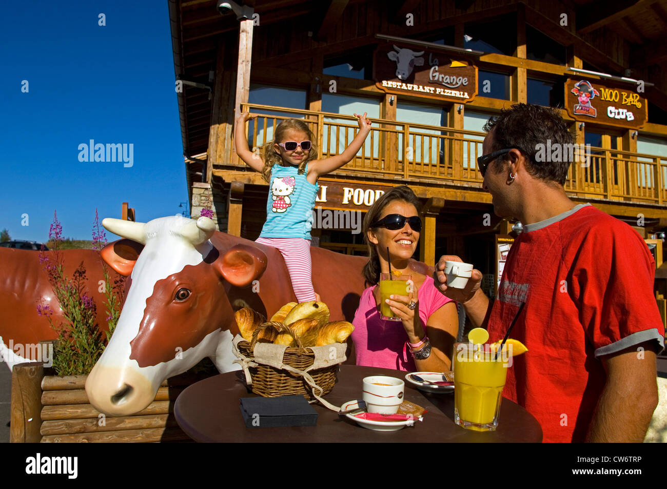 young family in front of mountain resort hotel, parents sitting at a cafe table, young daughter on a life-sized plastic cow, France, Savoie, Alps Stock Photo