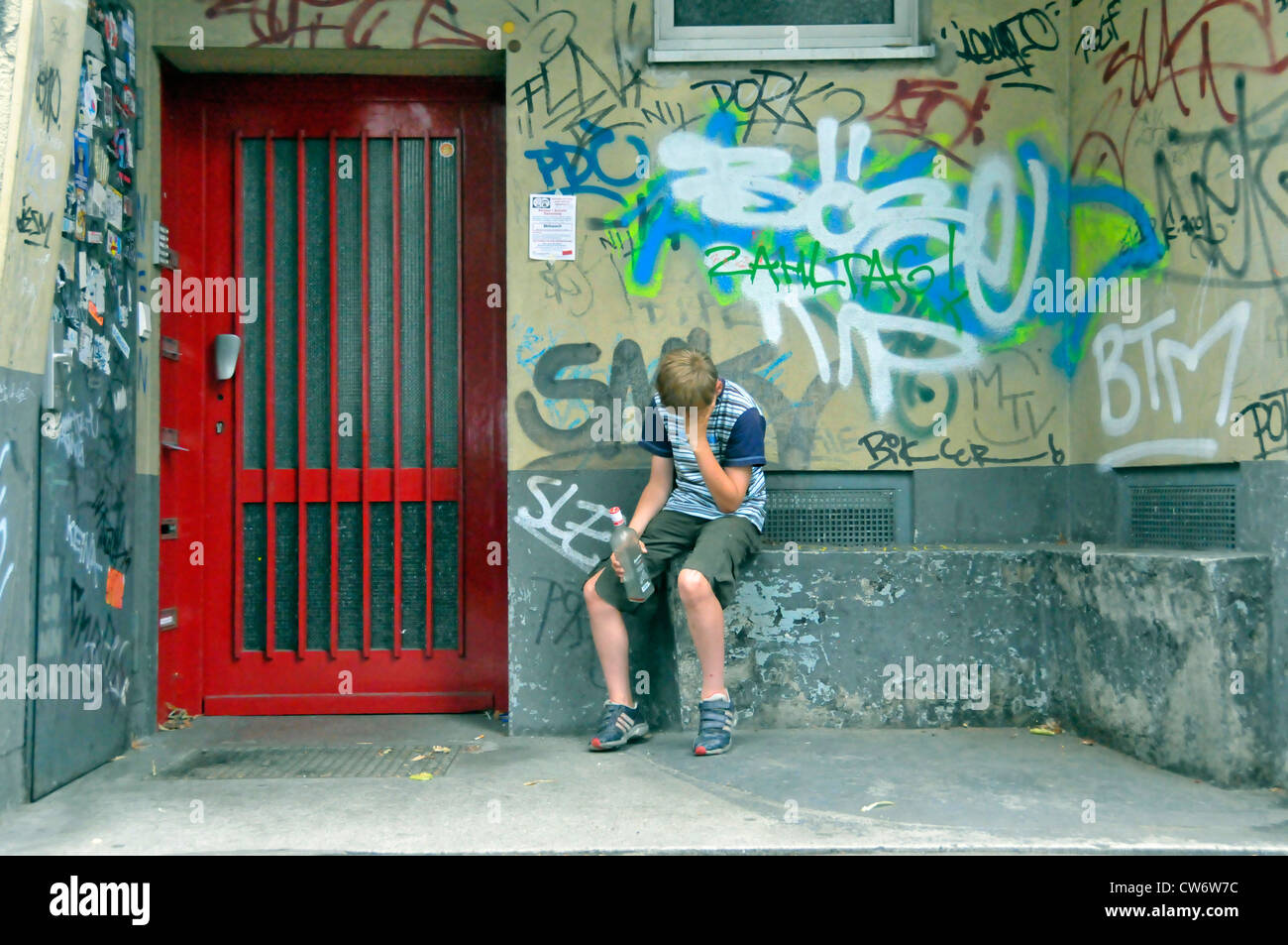 drunken teenager with alcohol bottle in front of a house entrance with graffitis, Germany, Cologne Stock Photo