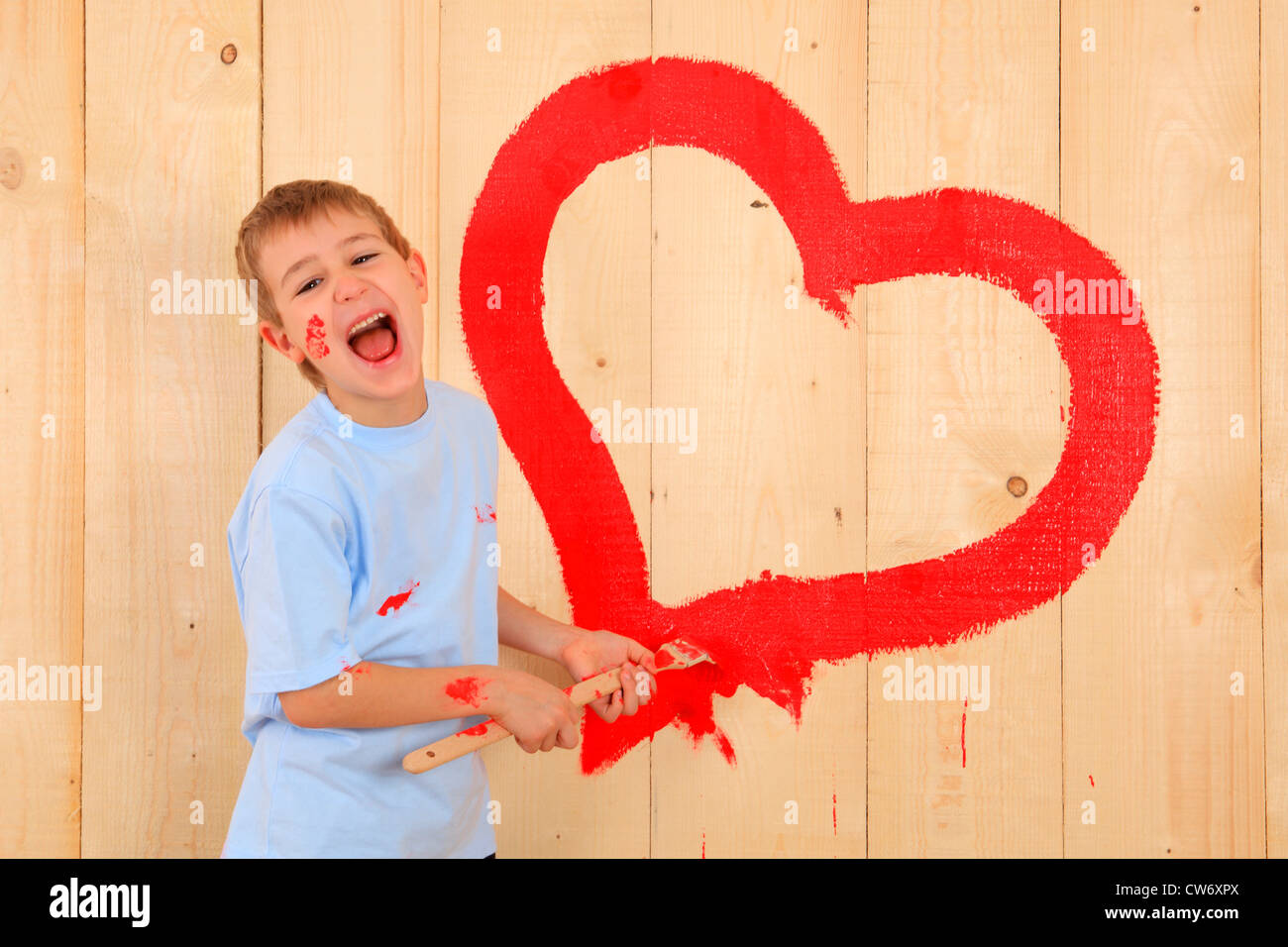 laughing boy painting a heart onto a wooden wall with red paint Stock Photo