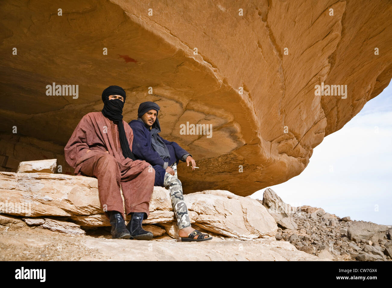 two Touaregs sitting under a rock spur at the Acacus Mountains showing a prehistoric rock painting, Libya, Sahara Stock Photo