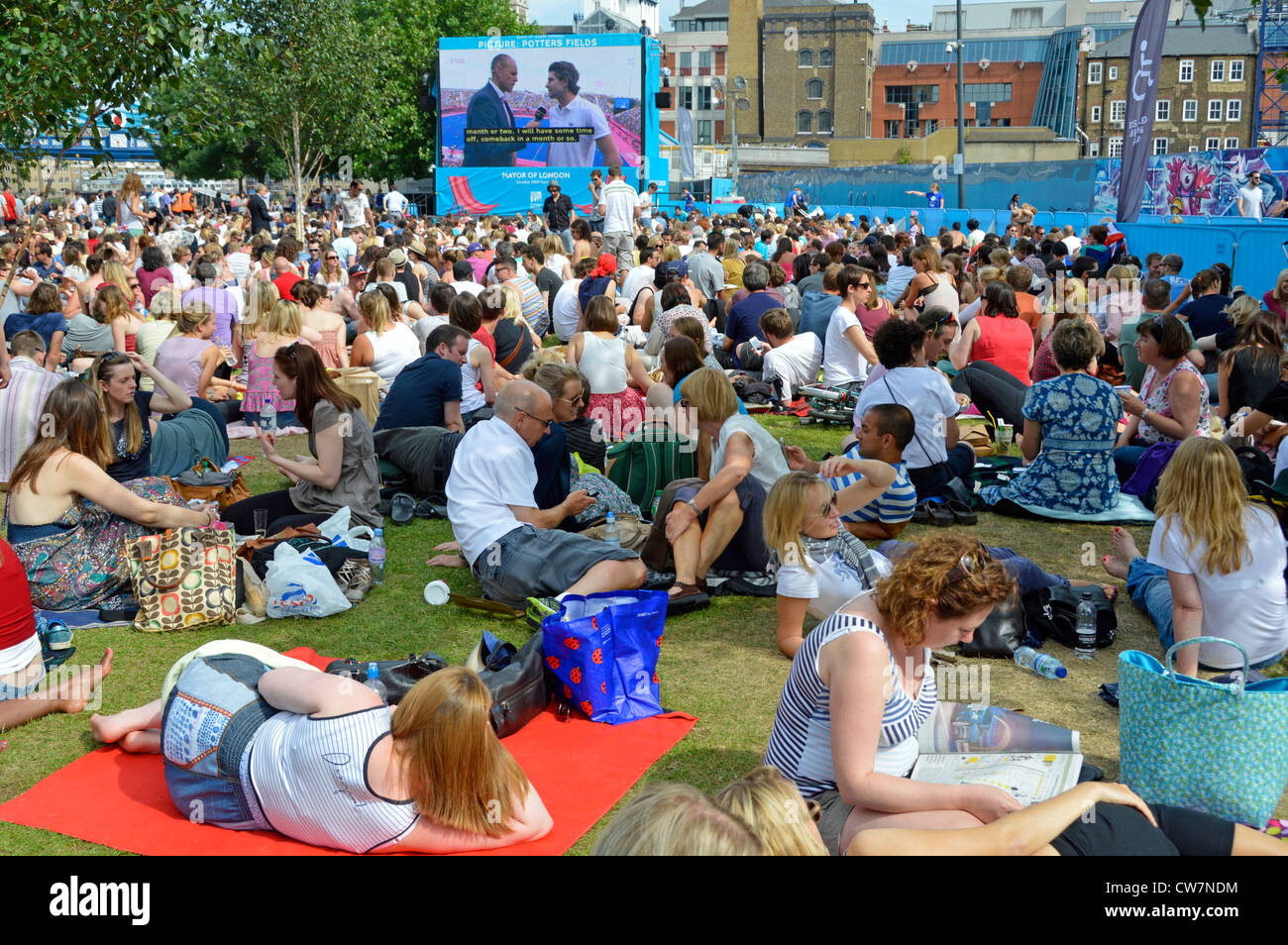 Crowd of people sit on grass watching London 2012 Olympics TV sport event large outdoor television screen in Potters Fields Park Southwark England UK Stock Photo