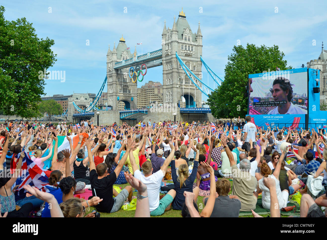 Crowd people sit on grass view London 2012 Olympic games TV large screen show hands up & waving Tower Bridge Potters Fields park Southwark England UK Stock Photo