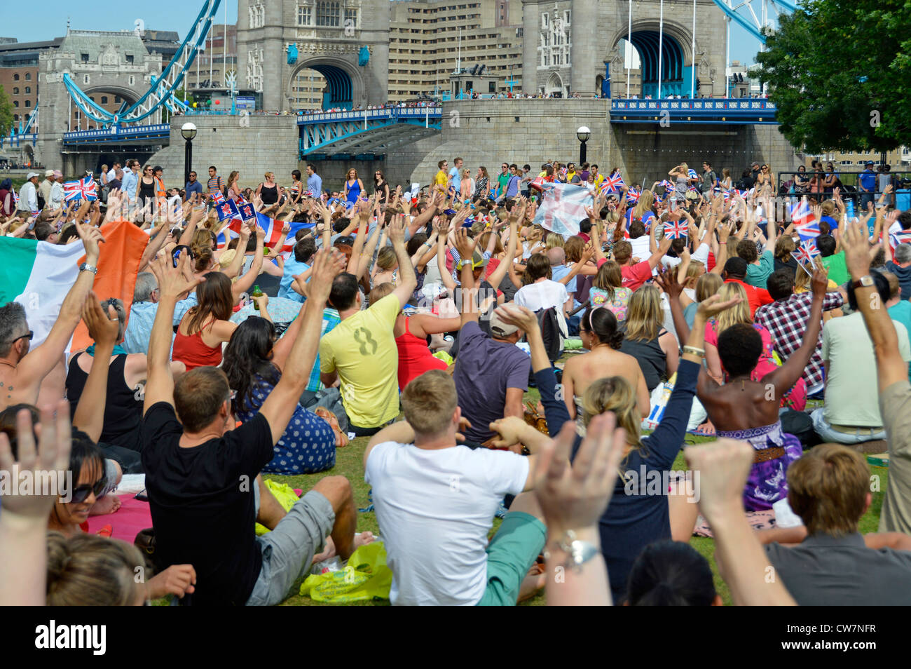 Crowd people sit on grass view London 2012 Olympic games TV large screen show hands up & waving Tower Bridge Potters Fields park Southwark England UK Stock Photo