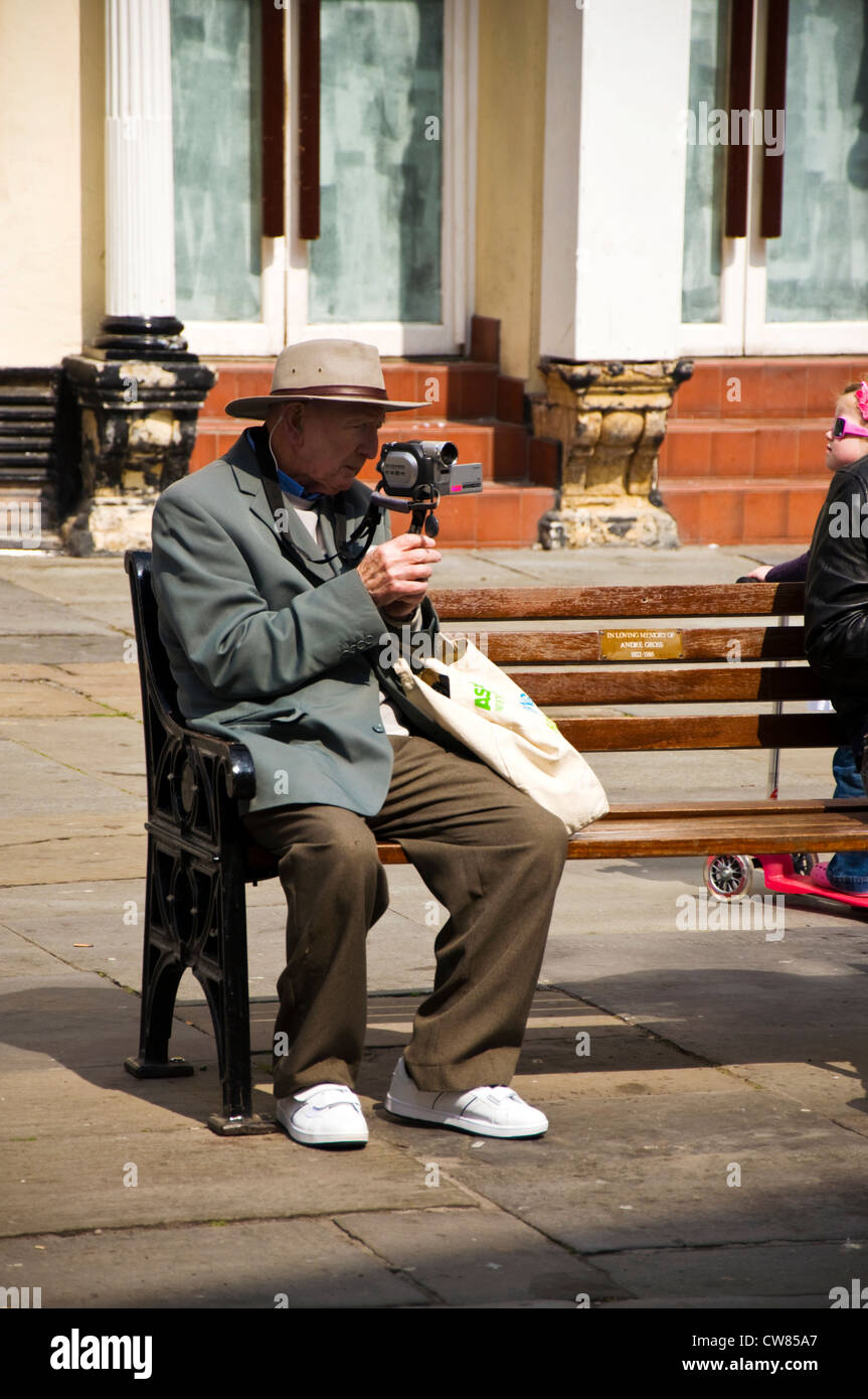 Senior man using a digital video camera sitting on a bench with white trainers sneakers in Bath Somerset England Stock Photo