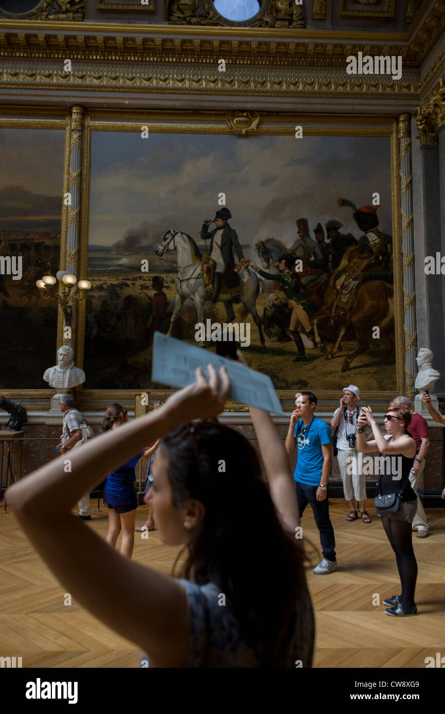 Tourists listen to guided audio tours and use cameras and smartphones to record art works below a painting of Napoleon at the Battle of Wagram, hanging in the Hall of Battles in the king's apartments of the Palace of Versaille, Paris. The Battle of Wagram (July 5–6, 1809) was one of the most important military engagements of the Napoleonic Wars and ended in a decisive victory for Emperor Napoleon I's French and Allied army against the Austrian. 'Napoleon at Wagram' was painted by Horace Vernet (Galerie des Batailles, Versailles). Stock Photo