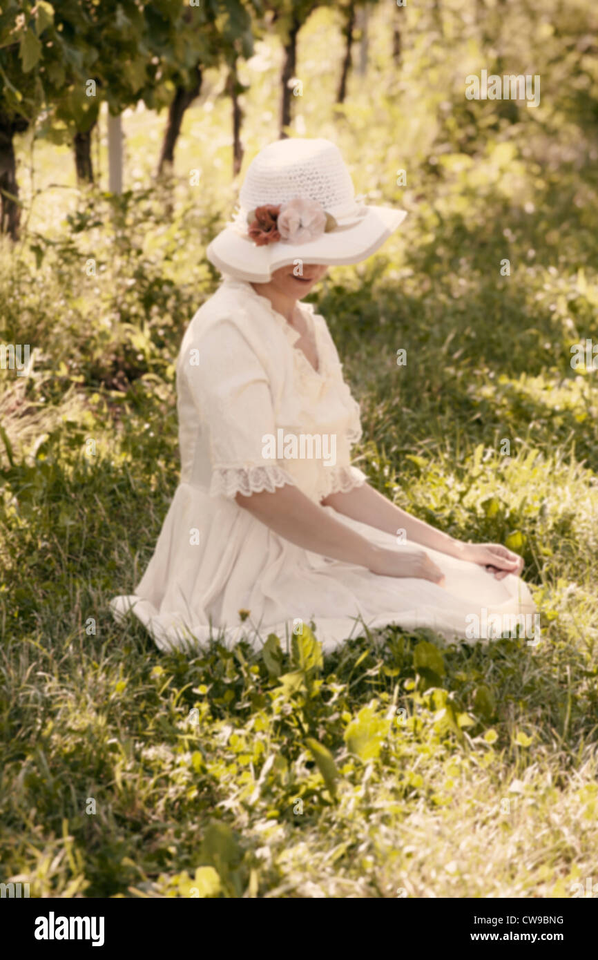 a woman in a white Victorian dress sitting on the grass between vines Stock Photo